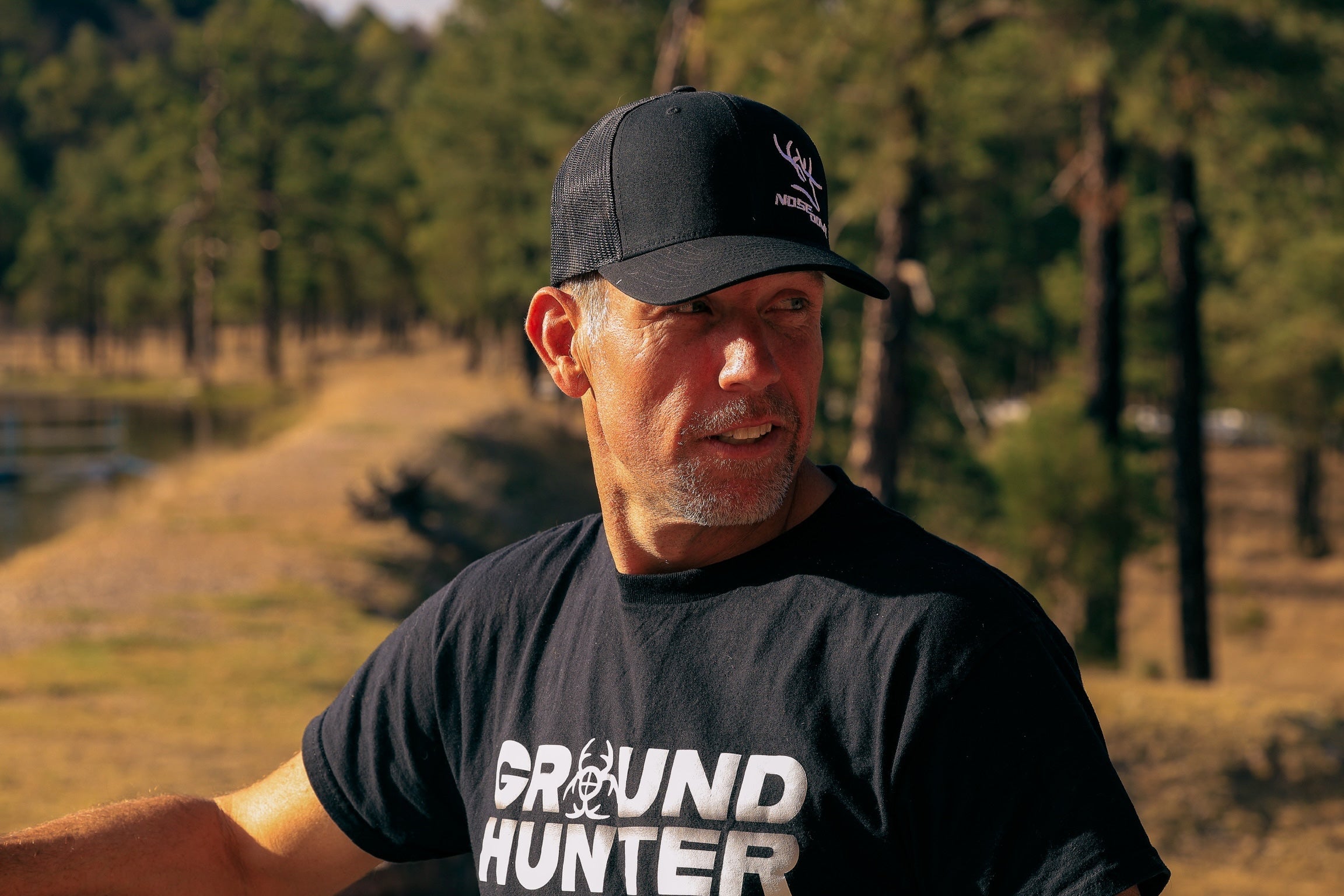 A man wearing a black baseball cap and a black Nukem t-shirt with 'GROUND HUNTER' printed in white, standing outdoors in a wooded area.